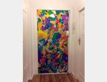 Tür in einem Innenraum, verziert mit einer bunten Collage aus farbigen Papierstücken. Umgeben von weißen Wänden und einem hellen Holzboden; rechts befindet sich eine Türklinke.