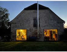 Ein altes, rustikales Gebäude mit einer Fassade aus verwittertem Mauerwerk, aufgenommen bei Dämmerung. Durch zwei erleuchtete Fenster sind ein gemütlich eingerichteter Innenraum und viele Pflanzen sichtbar; davor liegt eine grasbewachsene Fläche.