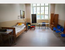 Ein heller Raum mit einem Pflegebett, einem Nachttisch und einem Hebebügel an der linken Wand. Im Raum befinden sich außerdem ein roter Couchtisch, ein Sofa, blaue Stühle, ein kleiner Tisch mit Stuhl, ein schwarzes Whiteboard und ein großer Fensterbereich mit viel Tageslicht.