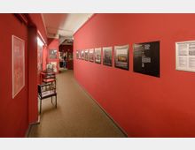 Ein langer, schmaler Gang mit roten Wänden, an denen gerahmte Poster und Fotografien hängen. Entlang der linken Wand stehen mehrere Stühle, der Boden ist mit Teppich ausgelegt, und am Ende des Gangs führt die Öffnung in einen weiteren Raum mit Menschen.