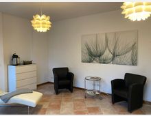 Ein minimalistischer Warteraum mit zwei schwarzen Sesseln, einem Glas-Beistelltisch und einem Liegestuhl. An der weißen Wand hängt ein großformatiges Bild mit botanischen Motiven, der Raum wird durch zwei moderne, blütenförmige Deckenlampen beleuchtet.