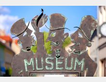 Metallkunstwerk mit drei stilisierten Figuren mit Hüten und Schnurrbärten, die das Wort „MUSEUM“ über einer gläsernen Öffnung tragen; im Hintergrund sind unscharfe Gebäude und blauer Himmel zu erkennen.
