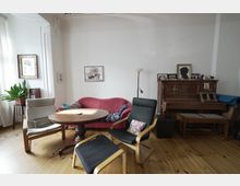 Wohnzimmer mit Holzboden, einem runden Holztisch in der Mitte und verschiedenen Sitzmöbeln, darunter ein rotes Sofa, ein schwarzer Sessel und ein Hocker. An der rechten Wand steht ein antikes Klavier mit Bildern und Dekorationen darauf, links stehen Pflanzen und Bilder an den Wänden.