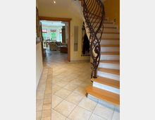 Ein Hausflur mit hell gefliesten Böden, einer Holztreppe mit geschwungenem, schmiedeeisernem Geländer auf der rechten Seite. Im Hintergrund öffnet sich ein Wohnbereich mit großen Fenstern, einem Sofa und Esstisch.