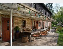 Hausfassade eines Backsteingebäudes mit überdachter Terrasse. Auf der Terrasse stehen eine lange Holztischgruppe mit Korbstühlen, farbige Lampen hängen von der Überdachung, und im Hintergrund ist ein Garten mit Blumen und Bäumen sichtbar.