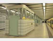 U-Bahn-Station mit gelbem Zug an einem Bahnsteig. Im Vordergrund befindet sich ein eckiges, grün-weißes Stationshäuschen mit Glasfenstern, umgeben von einer mit Fliesen verkleideten Wand.