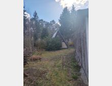 Kleiner Garten am Waldrand mit Rasenfläche und Sträuchern, im Hintergrund ein A‑Frame-Häuschen mit steilem Dreiecksdach. Rechts verläuft eine hohe Hauswand mit Kletterpflanzen; ringsum stehen hohe Nadelbäume unter blauem Himmel.