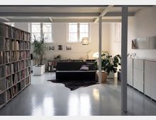 Großer, moderner Wohnraum mit weißen Wänden und einem grauen Boden. Links stehen hohe Bücherregale, in der Mitte befindet sich ein schwarzes Sofa auf einem dunklen Teppich, umgeben von Pflanzen und minimalistischen Möbelstücken. Große Fenster im Hintergrund lassen Tageslicht herein.