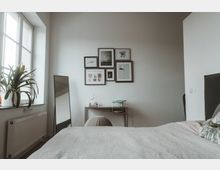 Minimalistisch eingerichtetes Schlafzimmer mit einem grauen Bett, einem Nachttisch und einem Wandspiegel. An der weißen Wand hängen mehrere Bilderrahmen, und ein großes Fenster mit einer Pflanze auf der Fensterbank sorgt für natürliches Licht.