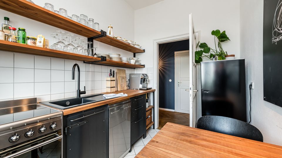 Helle, schmale Küche mit weißer Fliesenrückwand, Holzarbeitsplatte, schwarzer Spüle und offenem Wandregal mit Gläsern und Geschirr. Rechts steht ein schwarzer Kühlschrank neben einer Tür zum Flur; im Vordergrund ist ein Holztisch zu sehen.