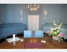 Ein kleiner, eleganter Saal mit blauen, gepolsterten Sitzbänken entlang der Wände und einer Holzkommode im Vordergrund, auf der Schreibutensilien und ein Blumenstrauß stehen. Im Hintergrund befindet sich eine weiße Tür, und eine moderne Deckenleuchte mit sichtbaren Glühbirnen hängt in der Mitte des Raumes.