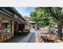Straßenszene mit einem Bistro auf der linken Seite, das mit Bildern von Speisen dekoriert ist, und Holzbänken im Außenbereich. Im Hintergrund sind ein Eingang zur U-Bahn-Station „Schlesisches Tor“, ein Backsteingebäude mit Arkaden und Bäume zu sehen, die teilweise Schatten spenden.