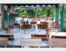 Eine Außenterrasse eines Restaurants mit Holzbänken und weißen Tischdecken. Auf den Tischen stehen kleine Blumenvasen und dekorative Kupferlichter; im Hintergrund sind Sonnenschirme und grüne Bäume zu sehen.