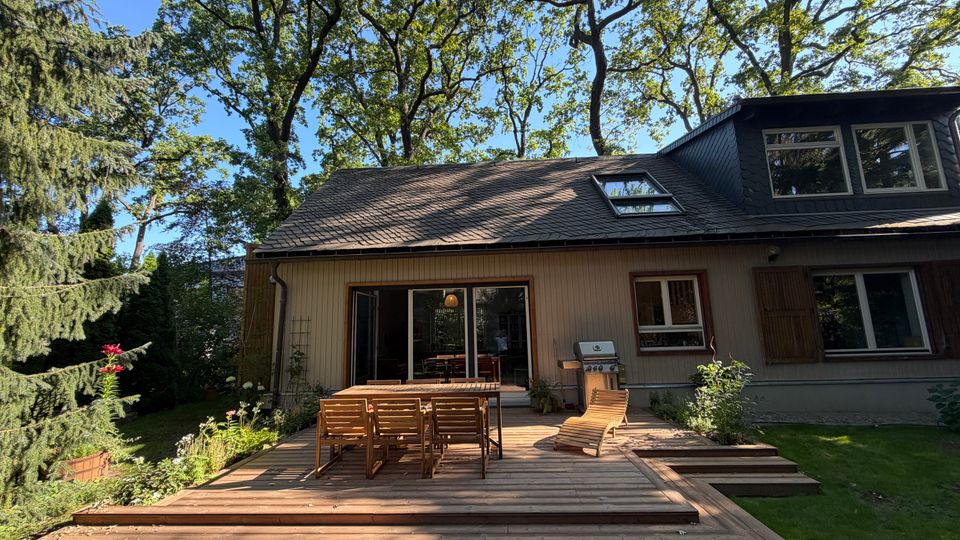 Holzterrasse an einem Einfamilienhaus mit geneigtem, dunklem Schieferdach, Dachfenster und großen Schiebetüren zur Gartenseite. Auf der Terrasse stehen ein Holztisch mit Stühlen, ein Grill und eine Liege; umgeben von Rasen und hohen Bäumen.