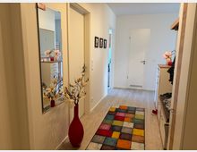 Heller, moderner Wohnungsflur mit hellem Holzboden, weißen Wänden und mehreren Türen entlang des Gangs. An der Wand hängen ein großer Spiegel und kleine Bilder; auf dem Boden liegt ein bunter Patchwork-Teppich, daneben steht eine rote Bodenvase mit künstlichen Blumen und rechts befindet sich eine Garderobe mit Kommode und Holzablage.