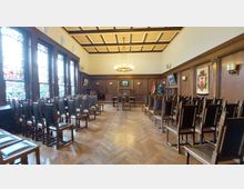 Innenansicht eines historischen Saals mit Holzwänden und Kassettendecke, dekoriert mit Wappen, Gemälden und farbigen Glasfenstern. Reihen von Holzstühlen sind auf einem Parkettboden angeordnet, und im Hintergrund befinden sich ein Tisch sowie Flaggen.