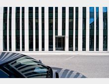 Moderne Gebäudefassade mit weiß-schwarzen vertikalen Streifen und großen Fensterflächen. Im Vordergrund ist ein geparktes Auto sichtbar, während die Gebäudeadresse "23" neben einem Eingang steht.