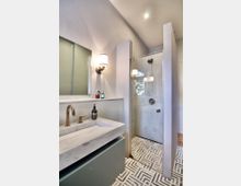 Modernes Badezimmer mit einem schlichten, rechteckigen Waschbecken aus weißem Marmor, einer bronzefarbenen Armatur und einer Duschkabine mit Glastür. Der Boden ist mit geometrisch gemusterten Fliesen ausgelegt, und die Wände sind in neutralen Tönen gehalten.