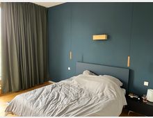 Ein Schlafzimmer mit einer dunkelblauen Wand, einem Bett mit weißer Bettwäsche und einem dunklen Kopfteil. Links befinden sich bodenlange, grüne Vorhänge, und an der Wand hängen minimalistische hölzerne Pendelleuchten und ein kleines Holzelement. Ein Nachttisch mit Gegenständen steht rechts neben dem Bett.
