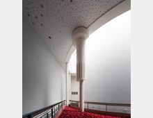 Ein moderner Innenraum mit hohen weißen Wänden, einer dekorativen Säule und einer durchgehenden Decke mit punktförmigem Muster. Der Boden ist mit rotem Teppich ausgelegt, der ein florales Muster hat, und ein Geländer mit Metallstreben grenzt den Bereich ab.