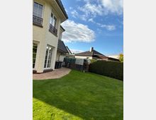 Einfamilienhaus mit hellem Putz und Fenstern mit dekorativen Schmiedeeisengeländern, links im Bild. Eine gepflasterte Terrasse und ein gepflegter Rasen erstrecken sich davor. Im Hintergrund sind weitere Häuser mit Schrägdächern und eine hohe Hecke sichtbar.