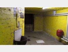 Ein schmaler Kellerraum mit gelb gestrichenen Backsteinwänden, freiliegenden Rohren an den Wänden und einer roten Druckkesselanlage. Im Vordergrund befindet sich ein Waschbecken mit einem Metalldeckel, der Boden ist aus Beton.