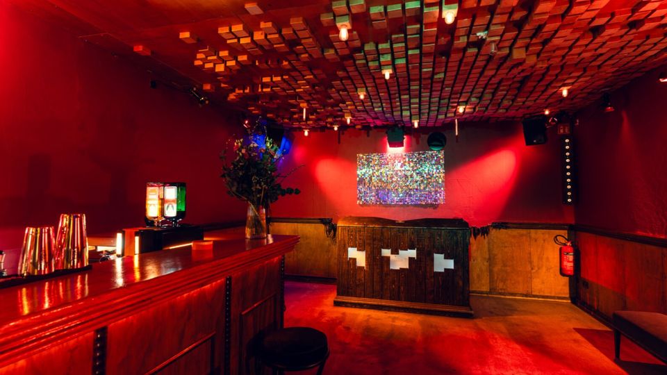 Innenraum einer Bar oder eines kleinen Clubs mit roten Wänden, Holztheke und Barhockern sowie einem DJ-Pult an der Rückwand. Auffällig sind die abgehängte Decke aus vielen Holzquadern, warme Deckenlampen und eine beleuchtete Wandfläche über dem Pult.