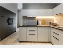 Moderne, L-förmige Einbauküche mit hellen Fronten, dunkler Arbeitsplatte und grauer Rückwand, beleuchtet durch eine durchgehende Unterbauleuchte. Links stehen hohe schwarze Schrankelemente mit integriertem Backofen; rechts sind Spüle, Toaster und Wasserkocher auf der Arbeitsfläche.