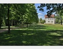 Ein Park mit einer Allee aus Bäumen, die Schatten auf die Rasenfläche werfen, daneben ein Kiesweg. Im Hintergrund steht ein weißes, zweistöckiges Gebäude mit rotem Dach, umgeben von weiteren Bäumen.