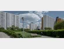 Wohnsiedlung mit mehreren modernen Hochhäusern in weißer und orangefarbener Fassadengestaltung, umgeben von Grünflächen, Gehwegen und einer Hecke. Im Vordergrund befinden sich eine Parkbank, eine Laterne und zwei Personen, die einen Weg entlanggehen.