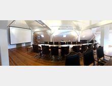 Ein heller Konferenzraum mit Dachschrägen, Holzboden und einer länglichen Tafel, die von lederbezogenen Stühlen umgeben ist. Im Hintergrund stehen ein Regal, eine Leinwand und eine kleine Buffetstation mit Getränken und Snacks.