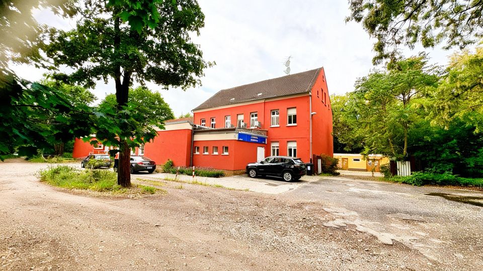 Zweigeschossiges, rot gestrichenes Gebäude mit Satteldach und mehreren Fenstern, davor ein kleiner Parkplatz mit Zufahrt. Das Haus liegt in einer grünen Umgebung mit großen Bäumen und dichtem Bewuchs am Rand des Geländes.