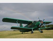 Grünes Doppeldecker-Flugzeug auf einer Wiese unter bewölktem Himmel mit einem Regenbogen im Hintergrund. Im Umfeld befinden sich flache Landschaft und ein Waldstreifen am Horizont.