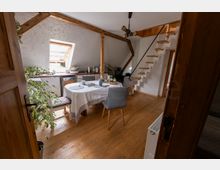 Heller Dachgeschossraum mit Holzboden, sichtbaren Deckenbalken und einem Esstisch mit weißer Tischdecke in der Mitte. Links eine kleine Küchenzeile unter einem Dachfenster, rechts eine schmale Holztreppe mit Geländer zu einer oberen Ebene.