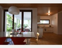 Offene Küche und Essbereich mit Holzdecke und großen Glastüren, die auf eine Terrasse mit Pflanzen und Sitzmöbeln führen. Der Raum ist minimalistisch eingerichtet, mit einem weißen Esstisch und roten sowie beigen Stühlen.