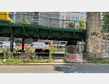 Eine grüne Stahlbrücke mit U-Bahn darauf überspannt eine Straße. Darunter befinden sich ungenutzte Bereiche mit Graffiti an den Betonpfeilern, wild wachsende Pflanzen und ein Zaun mit Plakaten; im Hintergrund sind geparkte Lieferwagen und Gebäude zu sehen.