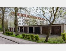 Eine Reihe einfacher Garagen mit Holztoren in einem Hinterhof, umgeben von Bäumen und Sträuchern. Im Hintergrund steht ein mehrstöckiges Wohngebäude mit weißer Fassade und roten Dachziegeln.