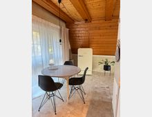 Kleine Esszimmer-Ecke mit Holzdachschrägen und natürlichem Licht. Ein runder, weißer Tisch mit drei schwarzen Stühlen steht auf einem hellen Steinboden, im Hintergrund ein cremefarbener Kühlschrank und eine Zimmerpflanze.