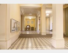 Heller Eingangsbereich eines historischen Gebäudes mit hohen Säulen, gelb gestrichenen Wänden und einer verzierten Kassettendecke. Der Boden besteht aus einem hell-dunkel karierten Muster, und an der Wand hängt ein Schaukasten mit Informationen.