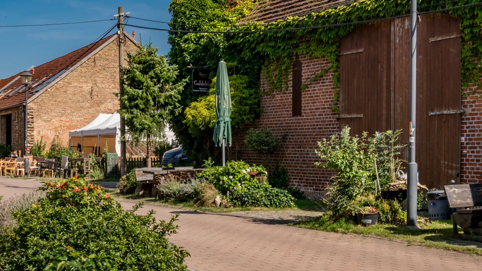 Dorfstraße mit Backsteingebäuden und großen hölzernen Scheunentoren, teils mit Efeu bewachsen. Am Straßenrand stehen Holztische und -bänke sowie ein geschlossener Sonnenschirm neben bepflanzten Beeten und Sträuchern.