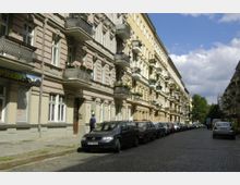 Straßenszene in einem Wohngebiet mit historisch anmutenden Gebäuden, die mehrere Stockwerke und verzierte Balkone haben. Entlang der gepflasterten Straße parken Autos, und einzelne Personen sind auf dem Gehweg zu sehen; im Hintergrund stehen Bäume und der Himmel ist teils bewölkt.