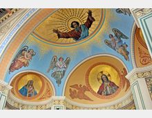 Deckenmalerei in einer Kirche mit christlichen Motiven, darunter Gottvater in einer goldenen Mandorla, Engel und Heiligendarstellungen auf blauem und goldverziertem Hintergrund. Verzierungen umfassen detaillierte Ornamente und Schriftzüge in den Bögen der Architektur.
