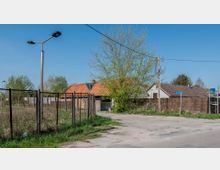 Ländlicher Straßenabschnitt mit einfachen Gebäuden, teils mit roten Ziegeldächern, umgeben von Zäunen und Bäumen. Die Umgebung wirkt ruhig, mit einer Mischung aus befestigten Flächen und Grasflächen.