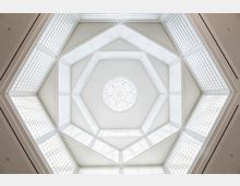Sechseckige Raumdecke mit einer zentralen, runden Kuppel aus weißem Glas und umgebenden Fensterelementen, die diffuses Licht hereinlassen. Die Struktur besteht aus geometrischen Formen und weißen Trägersäulen.