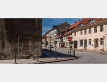Straßenecke in einer historischen Altstadt mit Kopfsteinpflaster und einer Reihe von Wohnhäusern mit Putzfassaden, teils Fachwerk und roten Ziegeldächern. Im Vordergrund stehen Verkehrsschilder und Poller mit Ketten, links dominiert eine hohe, fensterlose Brandwand.