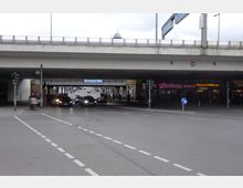 Straßenkreuzung mit mehrspuriger Unterführung und darüber verlaufendem Autobahnviadukt, auf dem Schilder mit Fahrtrichtungen angebracht sind. Im Hintergrund sind Geschäfte mit leuchtenden Werbeschildern sowie Fußwege sichtbar.