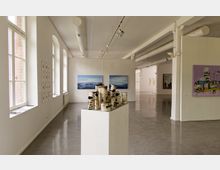 Heller Ausstellungsraum mit weiß gestrichenen Wänden und großen Fenstern auf der linken Seite, einer Skulptur aus Birkenstämmen auf einem Podest sowie mehreren Gemälden und Kunstwerken an den Wänden. Die Decke ist mit sichtbaren Rohren und Schienenstrahlern ausgestattet.