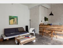 Minimalistisch eingerichtetetes Wohnzimmer mit einer Couch, einem weißen Stuhl und einem Couchtisch aus Holzpaletten. Rechts im Bild steht eine Bar aus Holzlatten mit zwei Hockern, darüber hängt eine schwarze Lampe; an der Wand hängen ein grünes Gemälde und ein Pflanzenarrangement.