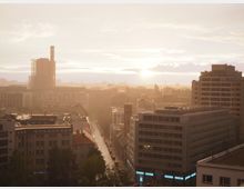 Städtische Skyline bei Sonnenuntergang mit modernen Bürogebäuden im Vordergrund und einem hohen Schornstein auf der linken Seite; eine Straße mit parkenden Autos verläuft durch die Szene, während die untergehende Sonne die Gebäude in warmes Licht taucht.