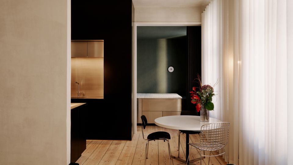 Modernes, minimalistisches Wohninterieur mit hellem Holzboden, einer schwarzen Küchenzeile mit Edelstahl-Nische und einem runden weißen Esstisch. Rechts sorgen bodentiefe, halbtransparente Vorhänge für Tageslicht; im Hintergrund ist eine Schlafnische mit Bett und dunkler Akzentwand zu sehen.